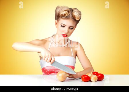 Young blonde housewife chopping onion on table in kitchen. Retro classic 50s style photoshoot. Stock Photo