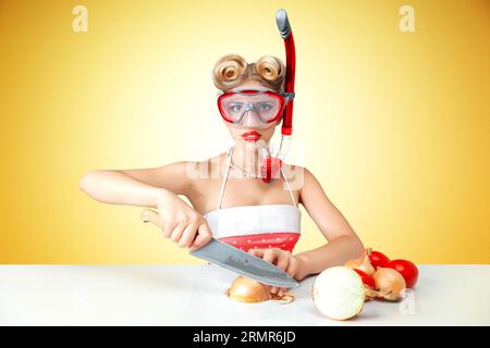 Young blonde housewife chopping onion in diving mask to protect her eyes. Retro classic 50s style photoshoot. Stock Photo
