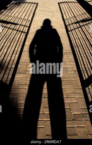 Foreboding shadow of person, legs apart, standing between two semi-open, wrought iron gates Stock Photo