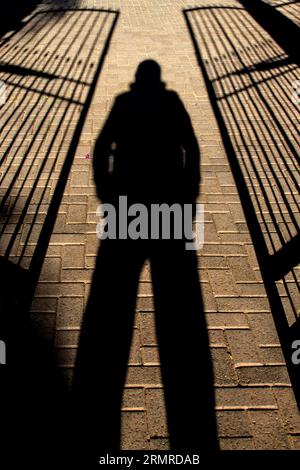 Foreboding shadow of person, legs apart, standing between two semi-open, wrought iron gates Stock Photo