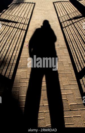 Foreboding shadow of person holding a phone to the ear, standing between two semi-open, wrought iron gates Stock Photo