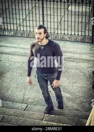 From above of muscular male with dark long hair in casual clothes looking up while climbing stairs on city street Stock Photo