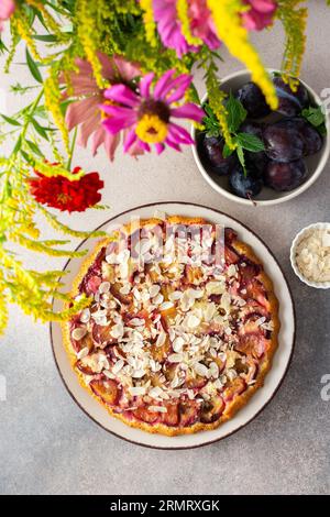 Plum cake with almond flakes, top view on the table with dessert and a vase of flowers, summer cake with seasonal fruits Stock Photo