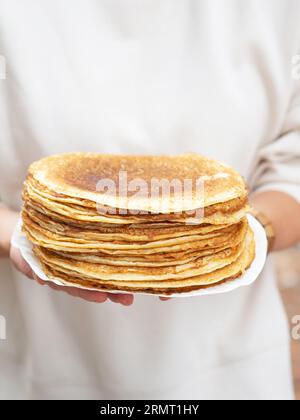 Women holding white plate with pile thin pancakes. Many pancakes are stacked. Thin pancakes with crispy crust. Pancakes for breakfast and carnival. Fo Stock Photo