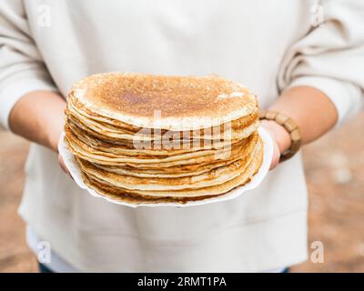 Women holding white plate with pile thin pancakes. Many pancakes are stacked. Thin pancakes with crispy crust. Pancakes for breakfast and carnival. Fo Stock Photo