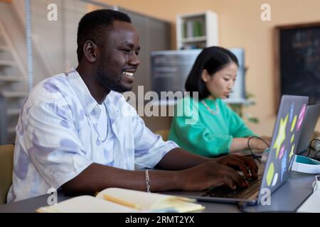 Smiling African American guy typing on laptop keyboard at informatics lesson while working over developing new software for decoding data Stock Photo