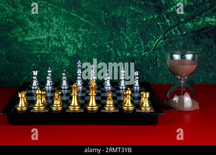 Chess board and silver and gold chess pieces with hourglass on a red surface and mottled background Stock Photo