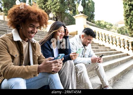 Three young diverse people texting with smartphones sitting on the stairs.Multiracial group smiling and using phones in a park sitting on a stone Stock Photo