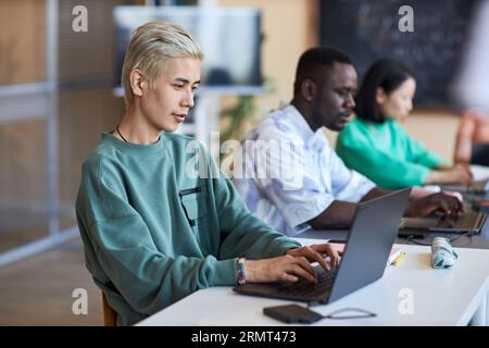 Side view of young classmates networking with focus on blond Asian guy typing on laptop keyboard while looking at screen and decoding data Stock Photo