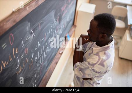 Pensive African American male student or teacher standing in front of blackboard with written formulas and equations in classroom Stock Photo