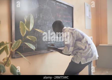 Young male student with tablet bending in front of blackboard while writing down formulas with chalk before explaining new subject Stock Photo