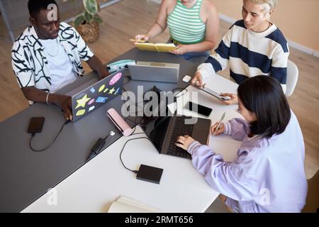 Asian female student using laptop and making notes in copybook at lesson of informatics while sitting in front of classmates with mobile gadgets Stock Photo