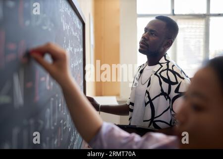 Young serious man in casualwear looking at equations on blackboard and thinking of ways of solving them at lesson of computer science Stock Photo