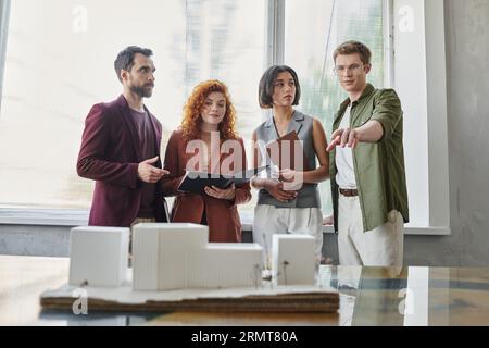 young architect pointing with hand at building model near investors in office, project presentation Stock Photo