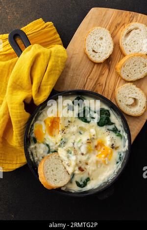 Healthy breakfasts or dinners, fried eggs with cheese, spinach leaves and coconut milk in a cast-iron pan, sliced baguette on a cutting board Stock Photo