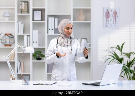 Young experienced female doctor in hijab using tablet computer for video call, female clinic worker consults patients remotely online, smiling contentedly inside clinic office. Stock Photo