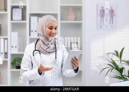 Young experienced female doctor in hijab using tablet computer for video call, female clinic worker consults patients remotely online, smiling contentedly inside clinic office. Stock Photo