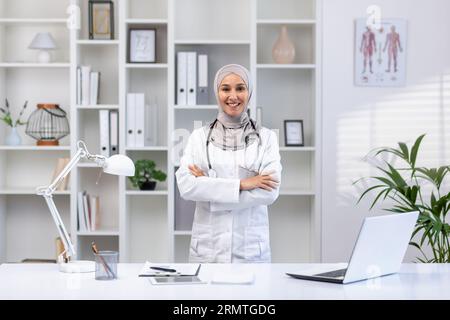 Portrait of young female doctor wearing hijab, Muslim woman smiling and looking at camera with arms crossed in white medical coat with stethoscope, working inside medical office office.. Stock Photo