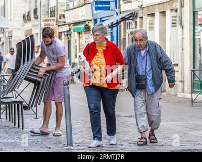 Elderly couple walking along cobbled street with cafe barman stacking chairs - Tours, Indre-et-Loire (37), France. Stock Photo