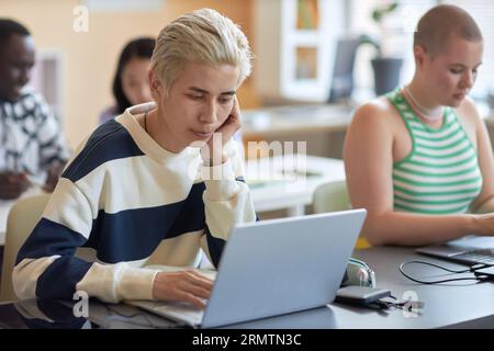 Young Asian blond man in pullover sitting in front of laptop screen while scrolling through coded data against his classmates at lesson Stock Photo