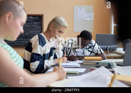 Focus on two young intercultural students carrying out examination test while sitting next to each other and writing down answers in copybooks Stock Photo