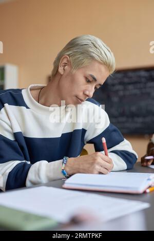 Young serious student in casualwear looking at copybook page while making notes or writing down answers to test questions at lesson Stock Photo