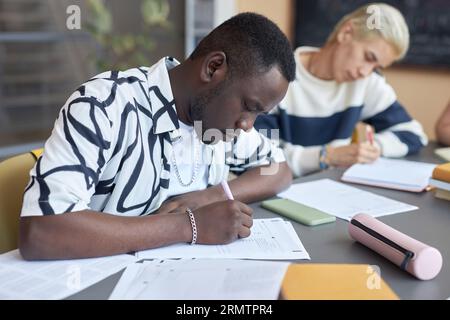 Serious African American guy making notes or putting ticks on paper while answering questions of examination test against Asian classmate Stock Photo