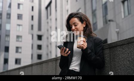 Close up. Caucasian female standing outdoors around grey modern buildings, working on her phone, smiling, looking around, and having drink from cup . Stock Photo