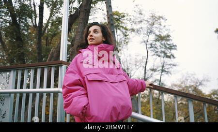 Pretty young woman going down stairs while walking in the park, holding on to the railing, wearing stylish pink jacket. Seasonal style, autumn season Stock Photo