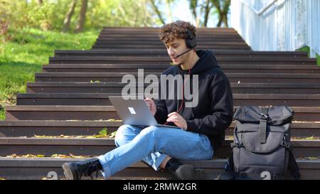 Smiling young man operator working with laptop while taking calls, using headset in the park. Autumn season. Education, working concept. Slow motion Stock Photo