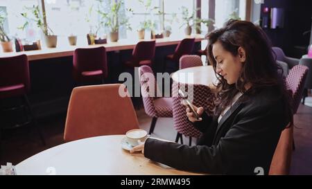 Caucasian girl drinking cappuccino inside of cafe, female is sitting at round table in the middle of the room and looking at the screen of her Stock Photo