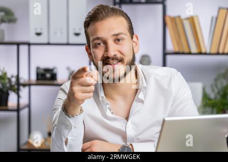 I choose you. Middle eastern businessman programmer software developer working on office laptop pointing to camera, looking with happy expression, making choice, showing direction. Arab freelancer man Stock Photo