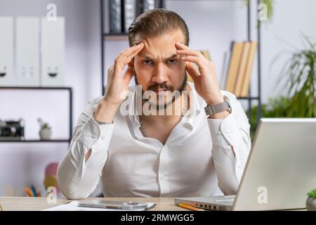 Tired ill middle eastern businessman suffering from headache problem tension migraine, stress at home office desk workplace. Bearded freelancer broker worker man works on laptop notebook. Overworking Stock Photo
