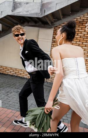 excited groom in sunglasses holding hands of african american bride and running on urban street Stock Photo