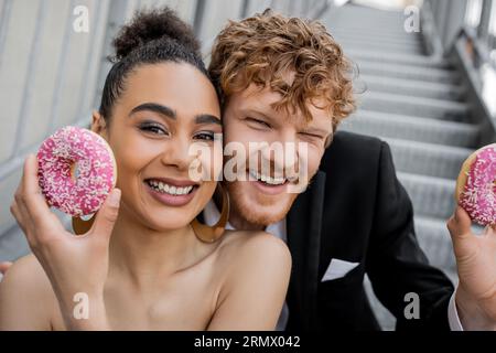 portrait of joyous multiethnic couple with donuts looking at camera, wedding on city street Stock Photo