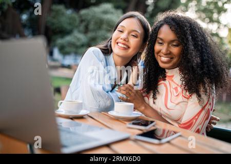 Portrait of happy young multiethnic female friends using laptop in cafe outdoors Stock Photo