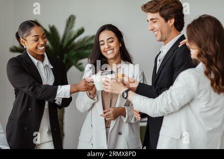 Happy multiracial male and female colleagues drinking coffee during break in office and clinking coffee cups Stock Photo