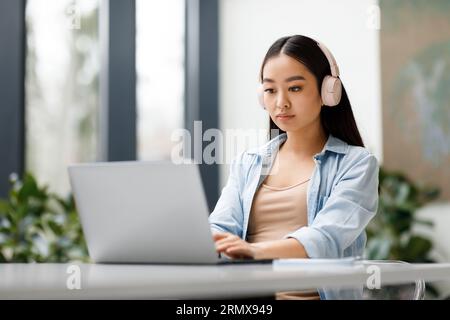 Asian lady student wearing headphones taking online class on laptop, sitting at table in university classroom Stock Photo