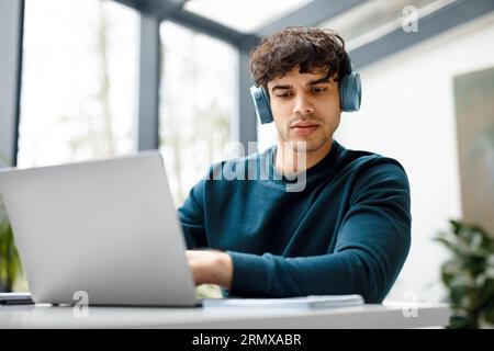 Focused guy student in wireless headphones using laptop and checking information in workbook, watching online tutorial Stock Photo