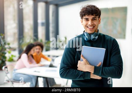 Happy student guy posing with notepads in hands and headphones on neck, smiling at camera while classmates studying Stock Photo