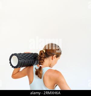 Conceptual photo of fitness, rear view of young athletic woman wearing sports bra holding foam roller on her shoulder. Stock Photo