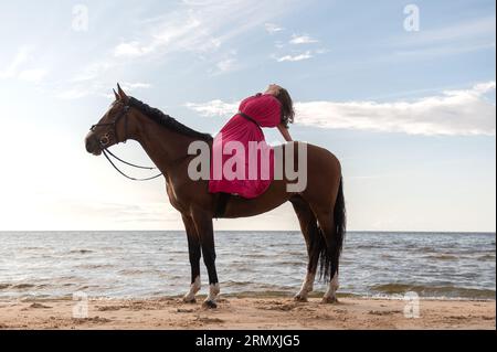 A youthful female wearing a pink dress posing on a horse on a beach Stock Photo