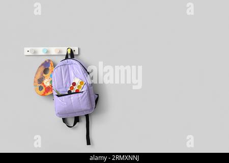 School backpack with paints and brushes hanging on light wall Stock Photo