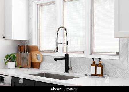 A kitchen sink detail with a herringbone tile backsplash, black and stainless steel faucet, white and black cabinets, and decor on the marble counter. Stock Photo