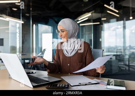 Young muslim businesswoman in hijab working in office with documents and talking on video call from laptop. Stock Photo