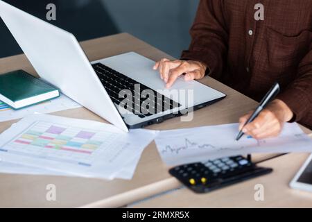 A close-up photo of the hands of a young woman working in the office at a desk, typing on a laptop and writing graphics and documents with a pen. Stock Photo