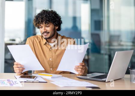 Successful hispanic financier on paperwork, young businessman investor checking papers, accounts contracts and statistical reports, man with laptop inside office smiling happy with achievement. Stock Photo