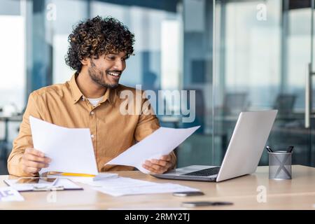 Successful hispanic financier on paperwork, young businessman investor checking papers, accounts contracts and statistical reports, man with laptop inside office smiling happy with achievement. Stock Photo
