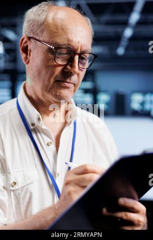 Old technician in cryptocurrency mining farm with specialized hardware using Blockchain technology. Fintech professional holding clipboard, inspecting asic rigs solving complex hashing operations Stock Photo