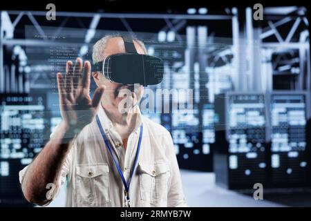 Octogenarian manager immersed in virtual reality at data center, doing equipment maintenance. Supervisor using VR headset to optimize server system performance, checking code lines Stock Photo
